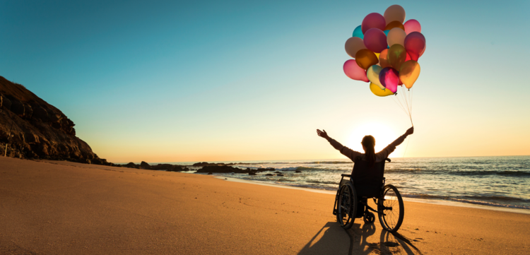 person in wheelchair on beach with upraised arms holding balloons