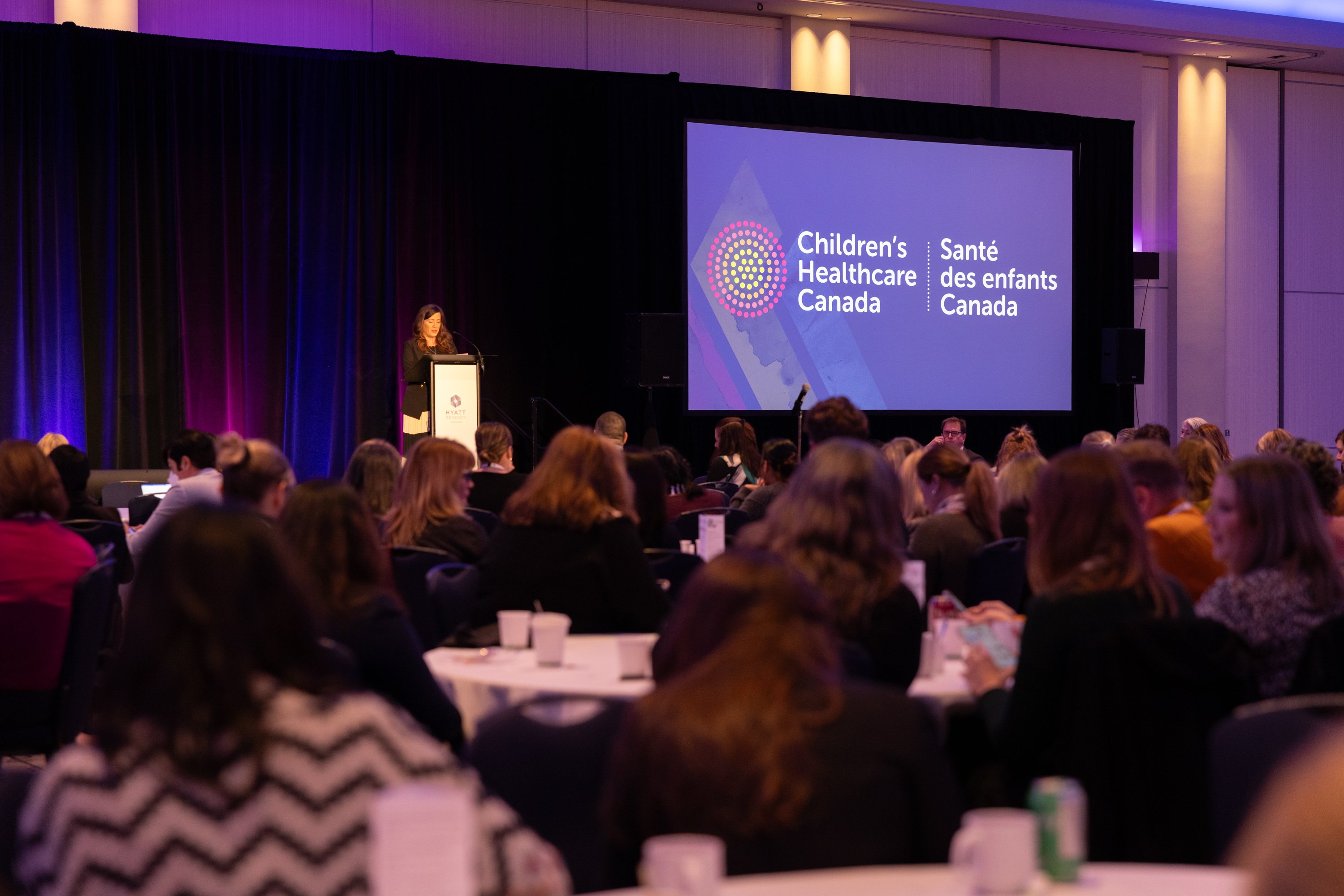 A photo of a speaker at a podium speaking to an audience. A large display screen is next to the speaker displaying the Children's Healthcare Canada logo. 