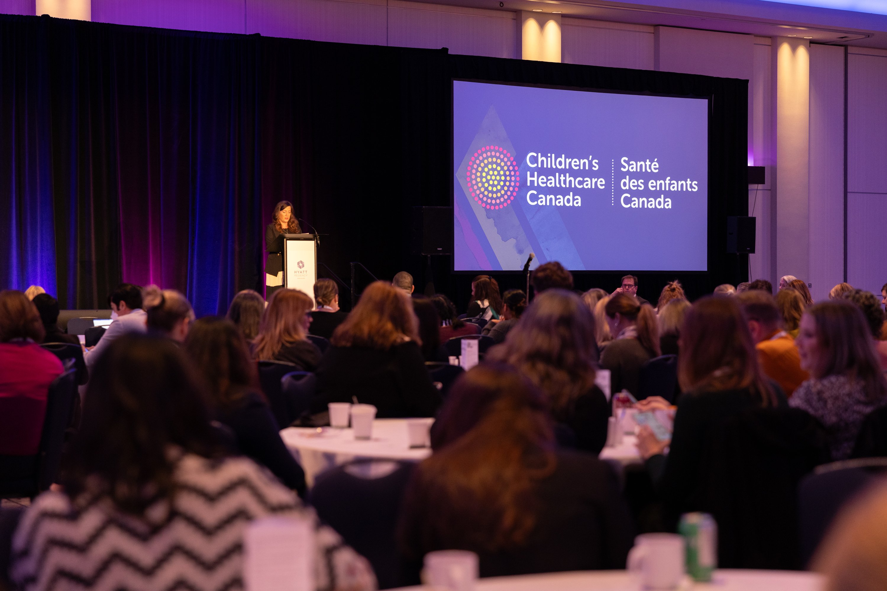 A photo of a speaker at a podium speaking to an audience. A large display screen is next to the speaker displaying the Children's Healthcare Canada logo.