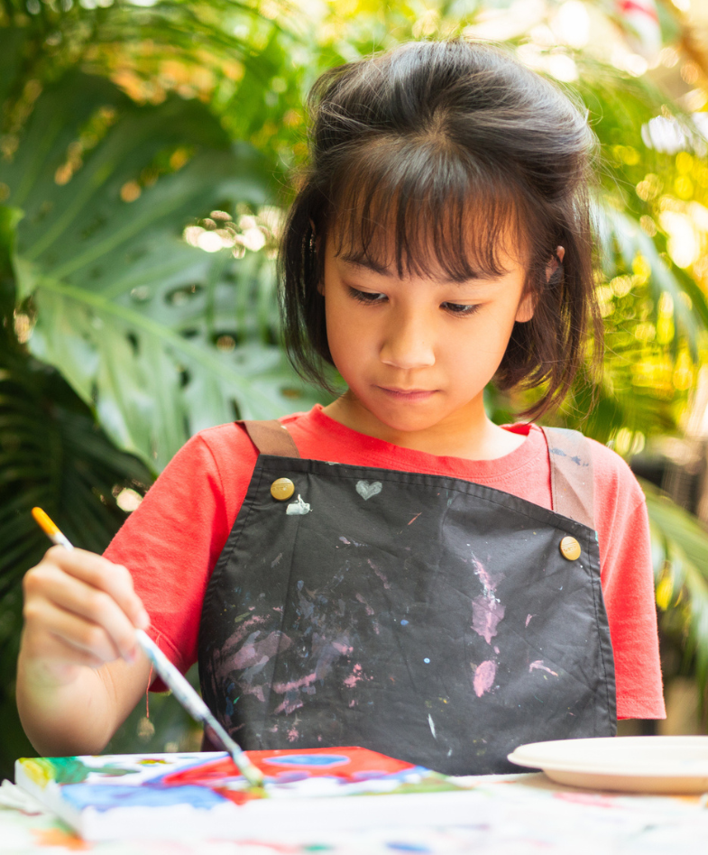 A young girl painting a picture in a red shirt and black smock 