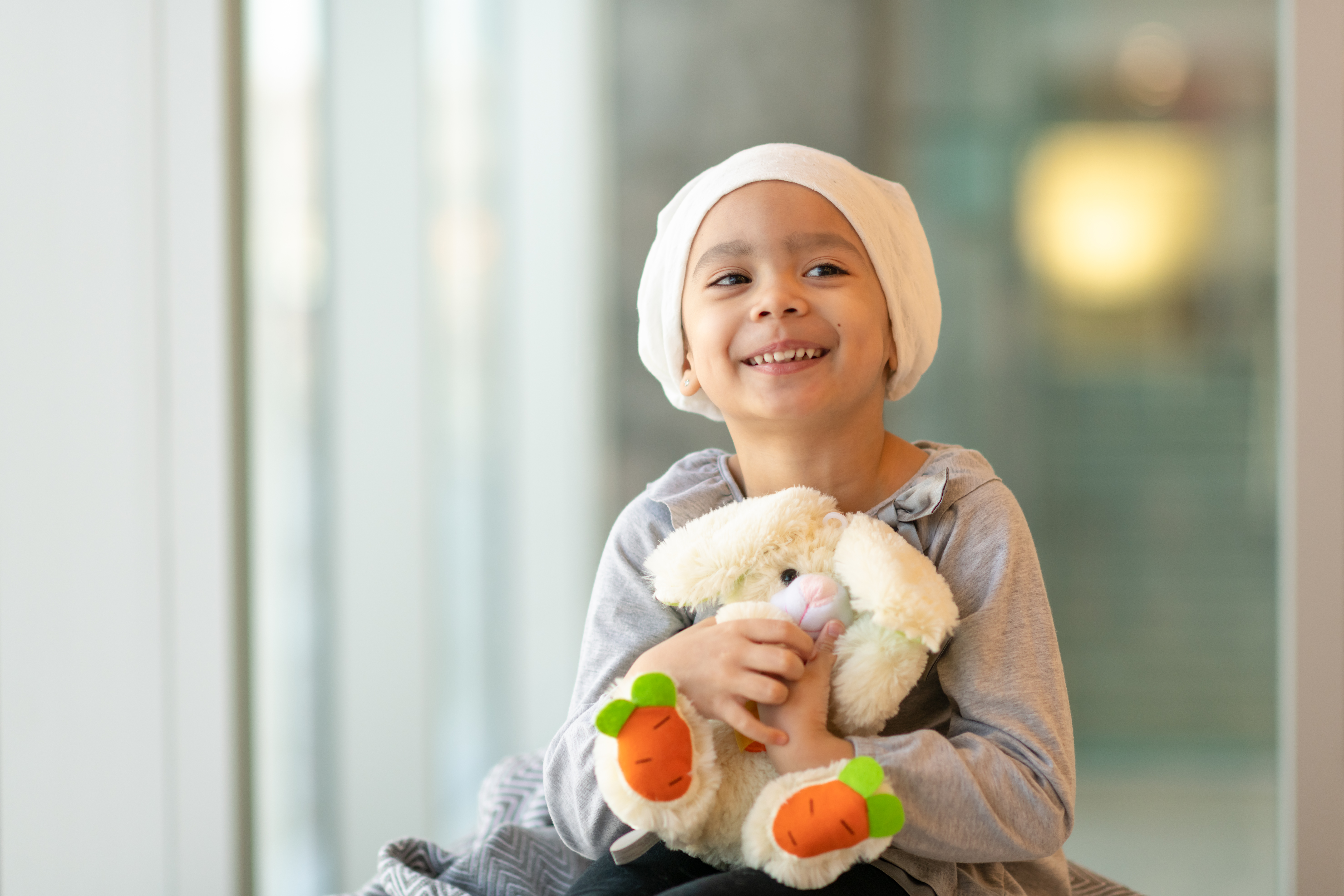 A little girl with cancer wearing a cream head wrap covering and clutching a stuffed bunny