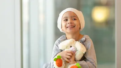 A little girl with cancer wearing a cream head wrap covering and clutching a stuffed bunny