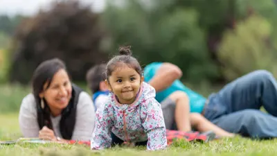 a young toddler girl crawling in front of her family lying down in the grass behind her